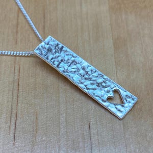 May include: A silver necklace with a hammered bar pendant featuring a cut-out heart shape.