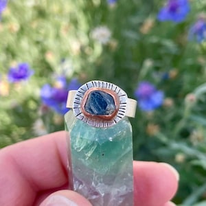 May include: A silver ring with a copper setting and a blue gemstone. The ring is set on a green crystal.