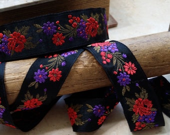 Puede incluir: Cinta negra con bordado floral rojo y morado. La cinta está envuelta alrededor de un carrete de madera.