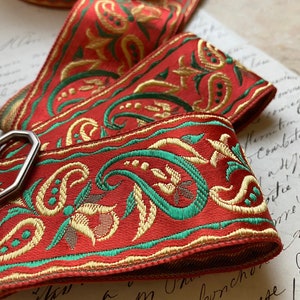 May include: Close-up of red ribbon with gold and green paisley and floral embroidery. The ribbon is folded over itself, showcasing the detailed design. The ribbon is likely used for crafting or decorative purposes.