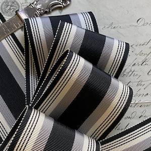 May include: Black and white striped ribbon with a silver and white stripe. The ribbon is folded and layered, showing the pattern.
