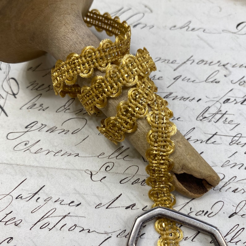 May include: Gold metallic trim with a woven design and scalloped edges. The trim is wrapped around a wooden spool.