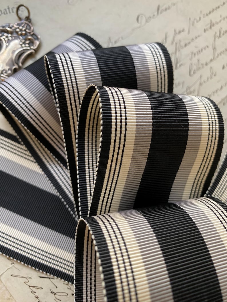 May include: Black and white striped ribbon with a cream and gray stripe. The ribbon is folded and has a textured surface.