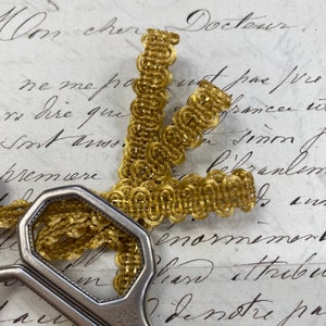 May include: Gold trim with a woven pattern, featuring a series of small circles. The trim is lying on a piece of paper with handwritten text.