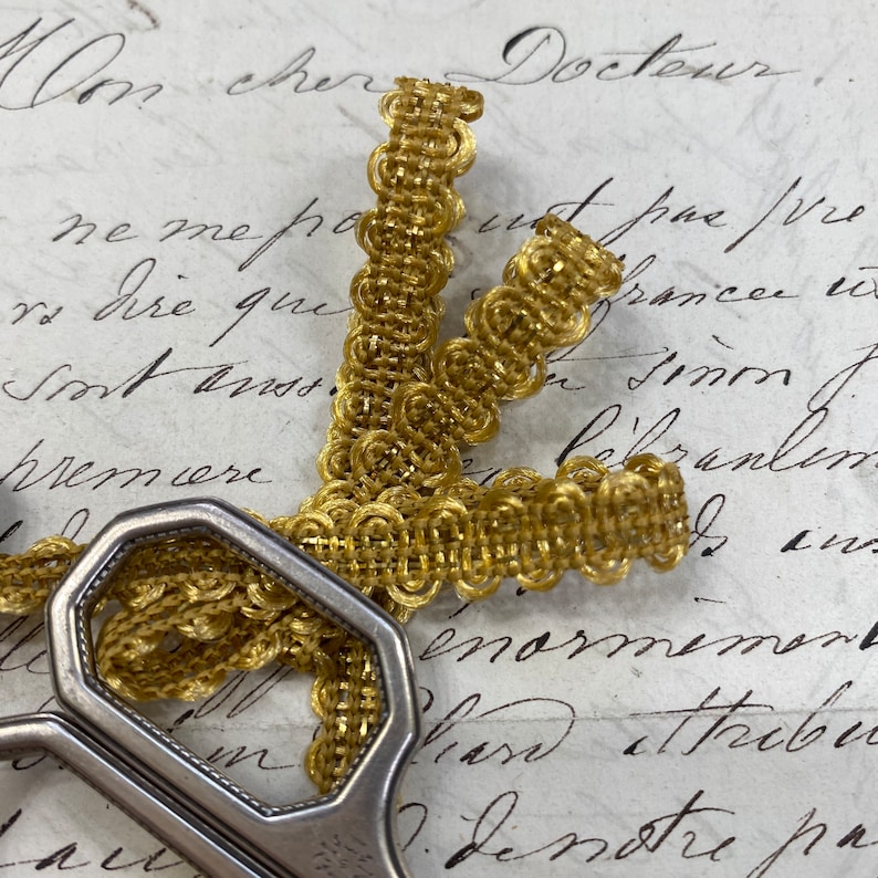 May include: Gold trim with a woven pattern, featuring a series of small circles. The trim is lying on a piece of paper with handwritten text.