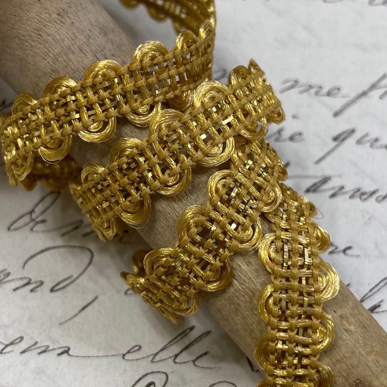May include: Gold metallic trim with a woven texture and scalloped edges. The trim has a delicate, intricate design.