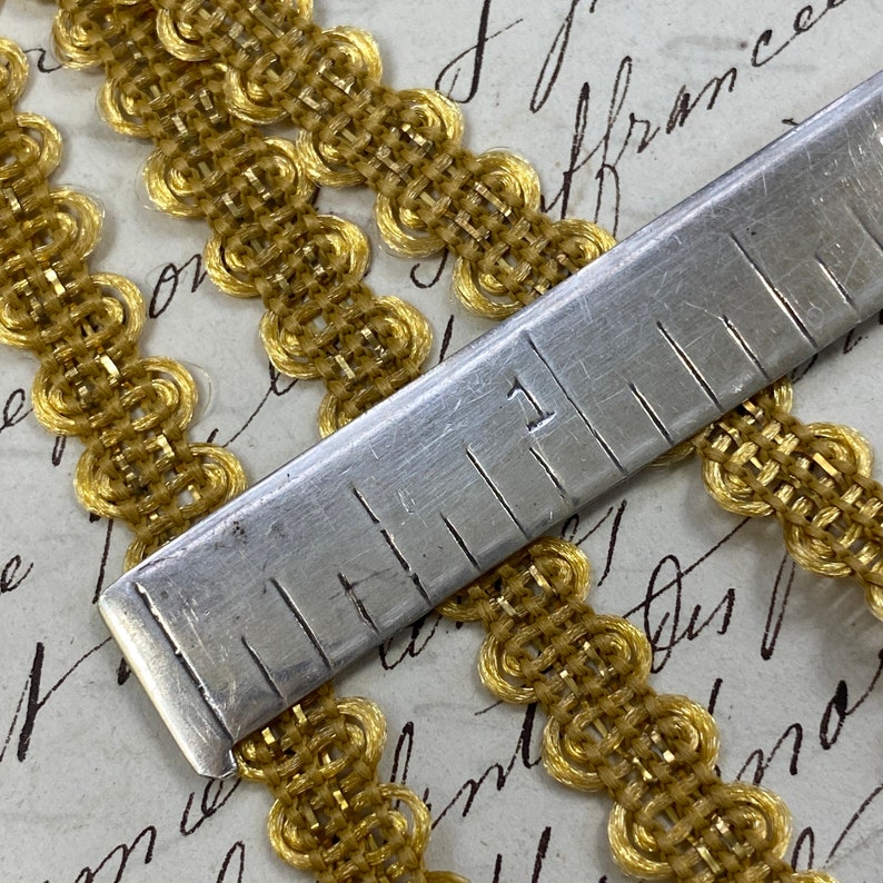 May include: Gold trim with a woven pattern and a repeating circle design. The trim is measured with a ruler, showing 1 inch.