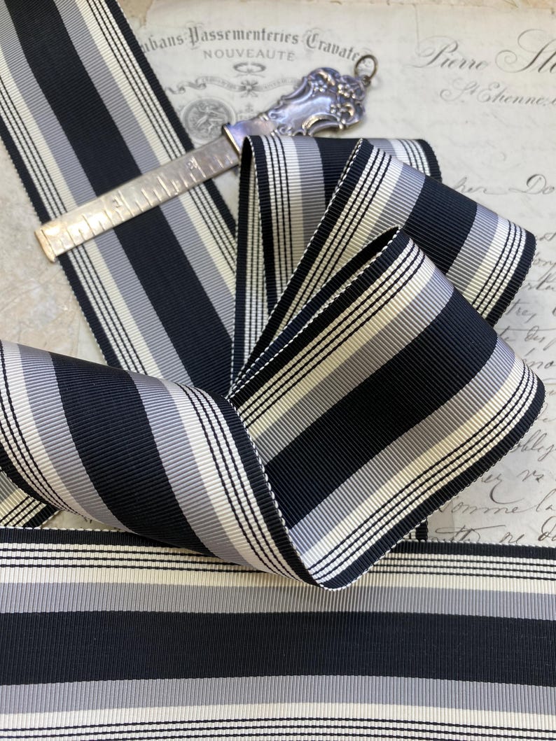May include: Black and white striped ribbon with a subtle gray stripe. The ribbon is folded and arranged on a vintage background.