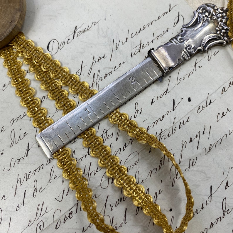 May include: A silver measuring tool with a decorative handle and a gold ribbon. The tool has markings for inches and centimetres.