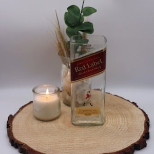 May include: A clear glass bottle with a red label that says "Johnnie Walker Red Label" and "Blended Scotch Whisky". The bottle is sitting on a wooden slice with a small candle in a glass jar.