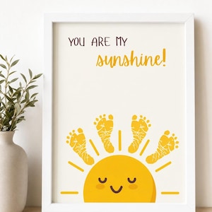 May include: White framed print with the text "YOU ARE MY sunshine!" above a yellow sun illustration. The sun is made of footprints. The print is on a light cream background. A vase with greenery is to the left of the print.