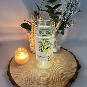 以下が含まれることがあります： 「Stolichnaya Vanilla Flavored Premium Vodka」とラベルが貼られた透明なガラス瓶。瓶は木の切り株の上に置かれ、隣には小さなろうそくがある。