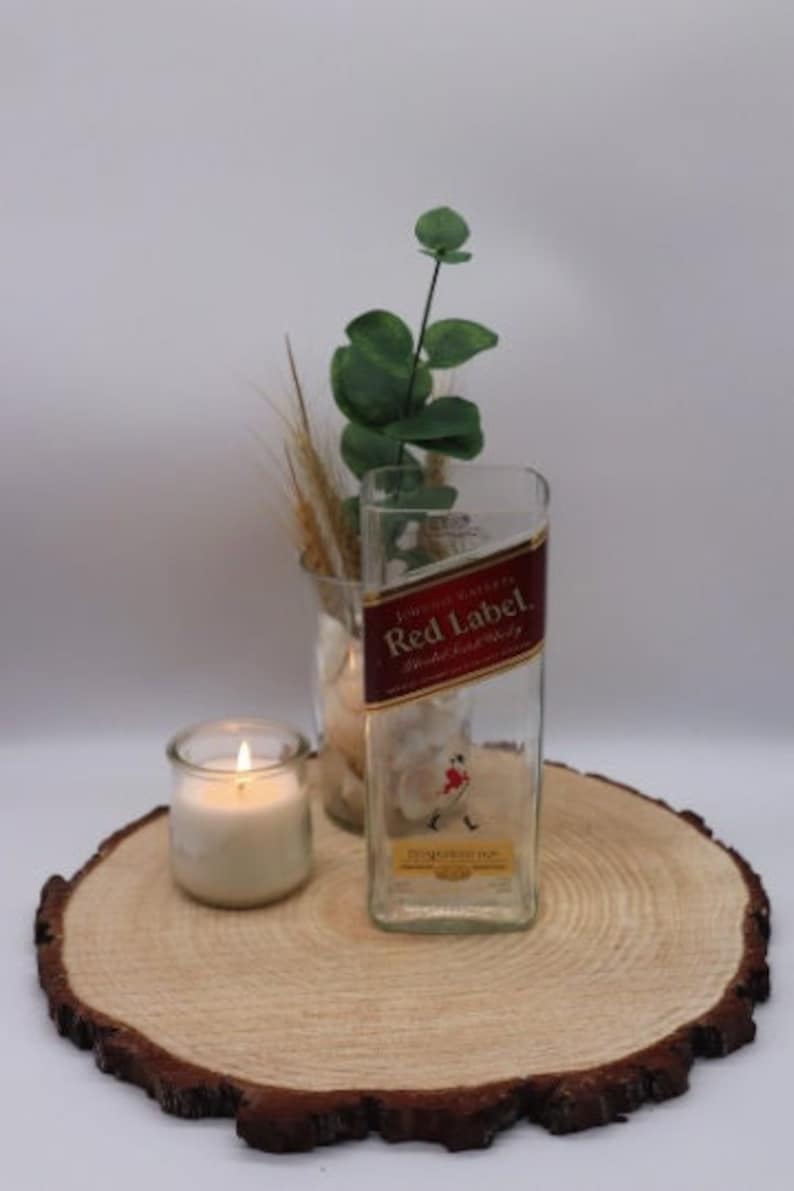 May include: A clear glass bottle with a red label that says "Johnnie Walker Red Label" and "Blended Scotch Whisky". The bottle is sitting on a wooden slice with a small candle in a glass jar.
