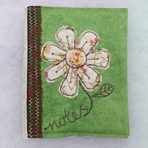 May include: A green fabric notebook cover featuring a brown spine with colourful decorative stitching. The front displays a large appliqued flower with a central button and the word notes embroidered in brown thread below the flower stem.