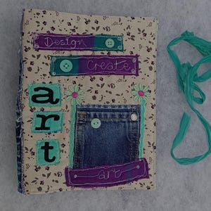 May include: A handmade journal with a floral fabric cover, denim pocket, and teal accents. The journal features the words "Design", "Create", and "art". A teal ribbon is tied to the side.