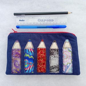 May include: A navy blue corduroy zippered pouch featuring five fabric pencil appliqués in various patterns. A black pencil, a blue pen, and a clear 15 cm Helix Oxford ruler are positioned above the pouch to provide a scale reference.