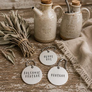 May include: Two speckled ceramic cruets with cork stoppers, one labeled OLIVE OIL and the other EXTRA VIRGIN OLIVE OIL. Three round silver metal tags on ball chains are inscribed with the words OLIVE OIL, BALSAMIC VINEGAR, and VINEGAR.