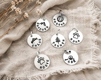 May include: Seven circular silver-toned metal tags, each featuring a unique engraved icon and a name. The icons include a mountain, basketball, tiara, pine tree, paw print, and peace sign. Each tag has a small metal jump ring attached at the top.