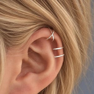 May include: A set of silver-toned ear cuffs consisting of one piece with a crisscross design positioned on the upper cartilage and a second piece featuring two parallel bands worn on the middle section of the ear cartilage.