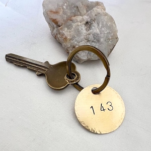 May include: A brass key and a round brass keychain tag with the number "143" stamped on it. The key and tag are connected by a brass ring. A rough, light-colored stone sits in the background.