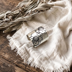May include: A wide silver cuff ring featuring a polished metallic finish and a stamped floral design. The surface of the ring displays two prominent flower motifs with textured centers and smooth, rounded petals, creating a distinct contrast.