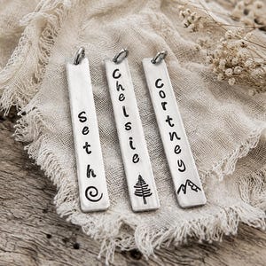 May include: Three rectangular silver-toned metal pendants with rounded corners and jump rings. Each pendant is vertically stamped with a name and a symbol: Seth with a spiral, Chelsie with a pine tree, and Cortney with a mountain range design.