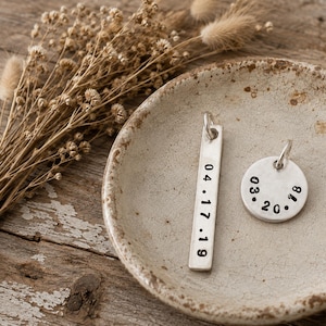 May include: Two silver-toned metal pendants, one rectangular and one circular, each featuring stamped black numbers. The rectangular pendant is stamped with 04.17.19 and the circular pendant is stamped with 03.20.18. Both pendants include a small jump ring.