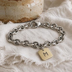 May include: A silver-toned chain link bracelet featuring a lobster claw clasp and a single square-shaped gold-toned pendant charm. The pendant is engraved with the uppercase letter M in a simple, clean font, centered on the flat metal surface.