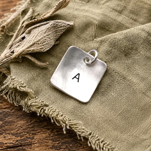 May include: A square silver pendant featuring the letter A in a black sans-serif font. The pendant has a brushed metallic finish and includes a small circular jump ring attached to the top corner for hanging on a chain or cord.