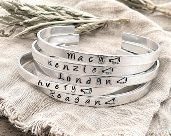 Puede incluir: Una pila de cinco brazaletes abiertos de metal plateado con acabado liso. Cada brazalete está grabado con un nombre único—Macy, Kenzie, Londyn, Avery o Reagan—seguido de un pequeño icono que representa un megáfono.