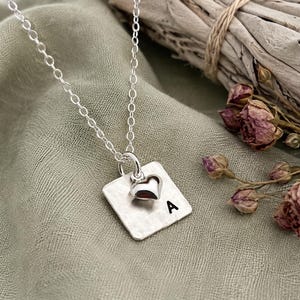 May include: A silver-colored necklace featuring a square pendant with a hammered texture and the letter A stamped in black. A small, polished silver-colored heart charm is attached to the same jump ring as the square pendant on a delicate chain.