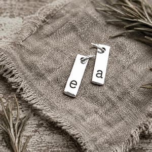 May include: Two rectangular silver-toned metal charms, each featuring a single lowercase letter stamped in black. One charm displays the letter e and the other displays the letter a. Both charms include a small circular jump ring at the top.