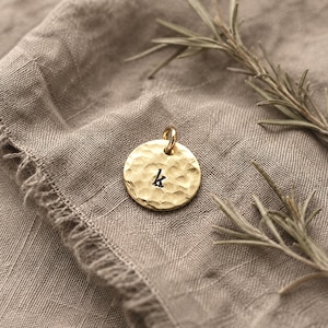 May include: A circular gold-toned pendant featuring a hammered, textured surface and the lowercase letter k stamped in black in the center. The pendant includes a small jump ring at the top for attachment to a chain or cord.