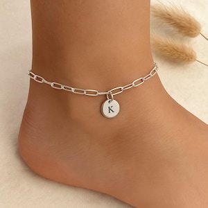 May include: A silver-colored anklet with a chain link design. A small, round charm with the letter "K" is suspended from the chain. The anklet is worn on an ankle.