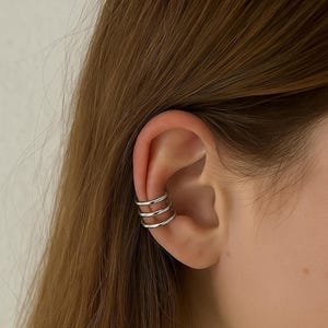 May include: Silver ear cuff featuring three stacked bands. The ear cuff is worn on the ear cartilage. The image shows the ear cuff on a person's ear with brown hair in the background. The jewelry is a simple, modern design.