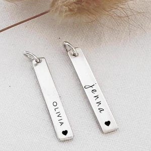 May include: Two silver bar pendants with a small heart at the bottom. One pendant says "Olivia" and the other says "Jenna".