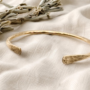May include: A gold-toned metal cuff bracelet featuring a hammered, textured surface. The bracelet has an open design with two rounded, flattened ends. The metal has a reflective, polished finish that catches the light, highlighting the indentations.