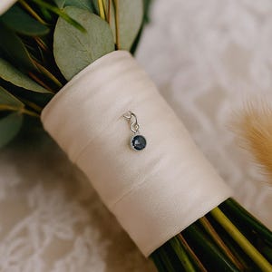 May include: A silver charm with a dark blue gemstone is attached to a bouquet of flowers. The charm hangs from a cream-colored ribbon wrapped around the stems of the flowers. Green leaves and stems are visible.