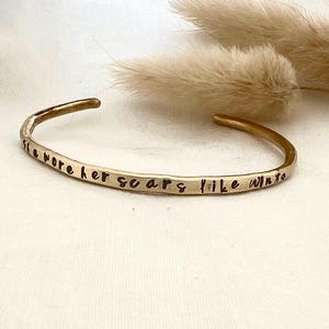 May include: A gold-colored cuff bracelet with the inscription "She wore her scars like wings." The open-ended bracelet has a hammered texture and is displayed on a white surface with dried floral accents.
