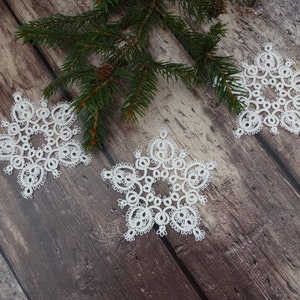 May include: Three white crocheted snowflakes on a brown wooden background. The snowflakes are delicate and intricate, with a circular design.