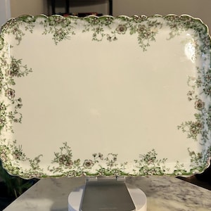 May include: A rectangular white ceramic serving tray with a scalloped edge and a decorative border featuring green leaves and small pink flowers. The center of the tray is plain white, and the rim is detailed with a delicate floral pattern.