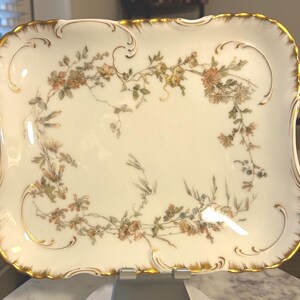 May include: A rectangular porcelain serving tray with scalloped edges and a gold-painted rim. The center features a delicate floral garland pattern in muted shades of pink, yellow, and green, arranged in a soft, flowing rectangular border.
