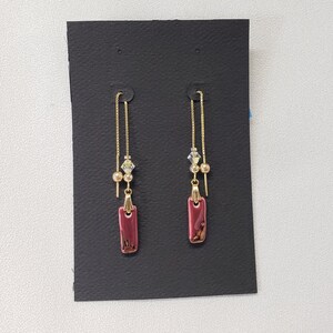 May include: A pair of gold dangle earrings with a burgundy rectangular charm and small gold beads and clear crystals.