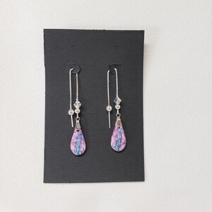 May include: A pair of dangle earrings with pink and blue teardrop-shaped glass beads. The beads have a marbled pattern and are attached to silver chains with small silver beads.