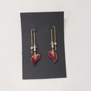 May include: A pair of dangle earrings with a red heart-shaped charm and gold chain. The earrings have a small gold bead and a clear crystal bead on the chain.