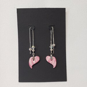 以下が含まれることがあります： ピンクのハート型のチャームとシルバーチェーンが付いた、ぶら下がりのイヤリングのペア。各チャームの上には、白と銀のビーズが付いています。