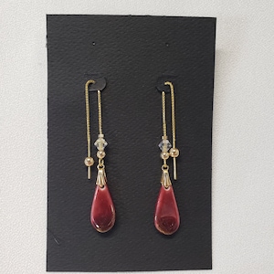 May include: A pair of dangle earrings with a burgundy teardrop-shaped ceramic charm and a gold chain. The earrings have a small clear crystal bead on the chain.