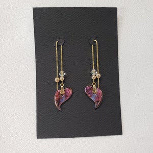 May include: A pair of dangle earrings with a purple heart-shaped charm and a gold chain. The earrings have a crystal bead and gold beads on the chain.