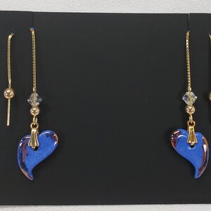 May include: A pair of blue heart-shaped dangle earrings with gold chains and beads. The earrings have a metallic finish and are on a black background.