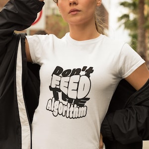 Może przedstawiać: Biała koszulka z napisem "Don't Feed The Algorithm" czarną, pogrubioną czcionką. Koszulka jest noszona przez osobę noszącą czarne okulary przeciwsłoneczne i czarną kurtkę.